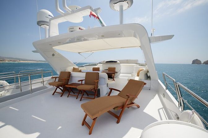 Sunlit yacht deck with brown cushioned wooden lounge chairs and shaded seating area overlooking calm blue ocean and distant rocky coastline
