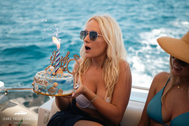 Woman in sunglasses on a yacht holding a seashell-decorated birthday cake with a lit sparkler, about to blow out a candle against the open ocean backdrop.