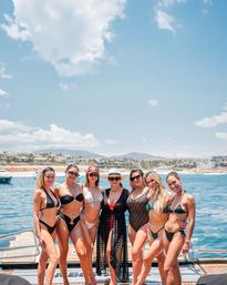 Seven women in bikinis posing on a yacht dock with turquoise ocean, sunny blue sky and a coastal marina with beachfront homes and hills in the background.
