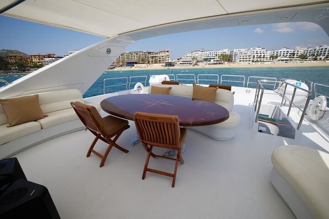 Sunlit luxury yacht deck with round teak dining table, wooden folding chairs and cushioned benches overlooking turquoise ocean and resort-lined coastline under a clear blue sky.