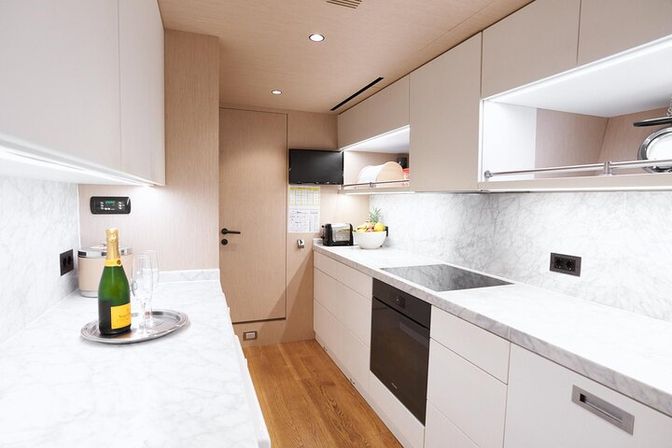 Compact modern galley kitchen with white marble countertops and backsplash, light wood cabinets, hardwood floor, built-in black oven and induction cooktop, champagne bottle and glasses on a silver tray, and a fruit bowl under cabinet lighting.
