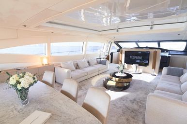 Bright cream-toned luxury yacht salon with panoramic ocean-view windows, skylight, plush sofas, round coffee table, marble dining table with floral centerpiece, and a flat-screen TV.