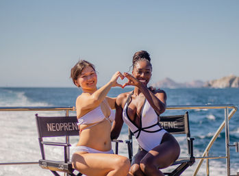 Two people in white swimsuits on a sunny boat deck at sea, sitting on deck chairs and smiling while making a heart shape with their hands against a coastal ocean backdrop.