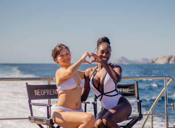 Two people in white swimsuits on a sunny boat deck at sea, sitting on deck chairs and smiling while making a heart shape with their hands against a coastal ocean backdrop.