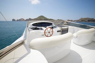 White luxury yacht helm with wooden steering wheel and plush white seating overlooking turquoise sea and rocky island coastline under a sunny blue sky