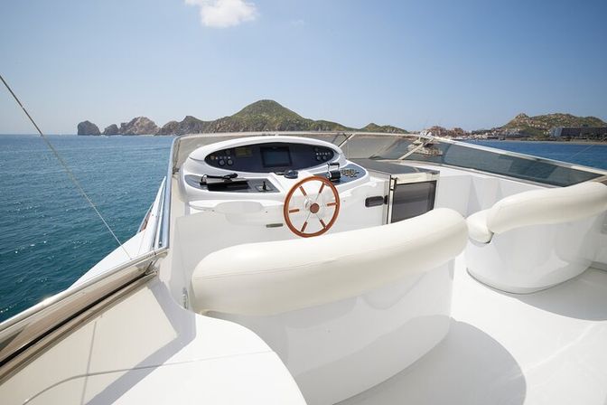 White luxury yacht helm with wooden steering wheel and plush white seating overlooking turquoise sea and rocky island coastline under a sunny blue sky