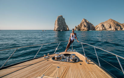Woman in a white swimsuit posing on the teak bow of a yacht with deep blue ocean and dramatic Cabo San Lucas rock formations rising under a clear sky.