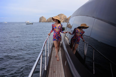 Woman in a colorful swimsuit cover-up and straw hat walking barefoot along a yacht’s wooden deck, her reflection on the glossy hull, with boats and rocky coastal arches on the ocean horizon.