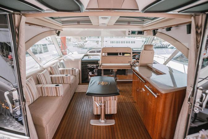 Luxury yacht interior salon with teak floors, striped cushioned bench seating, central captain’s chair and helm station, wood galley countertop and marina view through large windows.