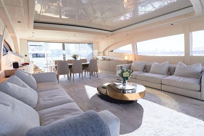 Sunlit luxury yacht salon with plush cream sofas and pillows, round gold-and-glass coffee table topped with fresh flowers, dining table and chairs, and panoramic windows framing a marina and distant city skyline.