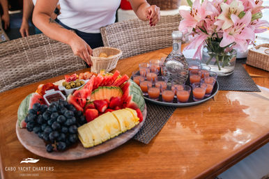 Vibrant tropical fruit platter (pineapple, watermelon, grapes, strawberries) and small bites beside a tray of peach-colored cocktail shots and a decanter on a wooden yacht table, pink lilies nearby and a hand reaching for crackers.