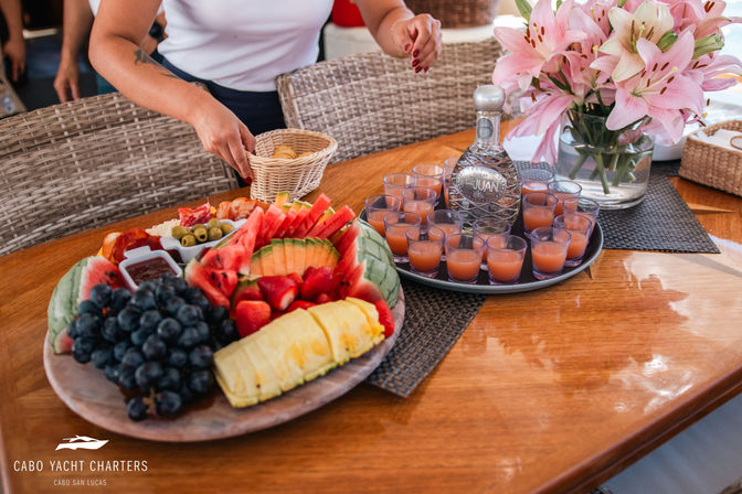 Vibrant tropical fruit platter (pineapple, watermelon, grapes, strawberries) and small bites beside a tray of peach-colored cocktail shots and a decanter on a wooden yacht table, pink lilies nearby and a hand reaching for crackers.