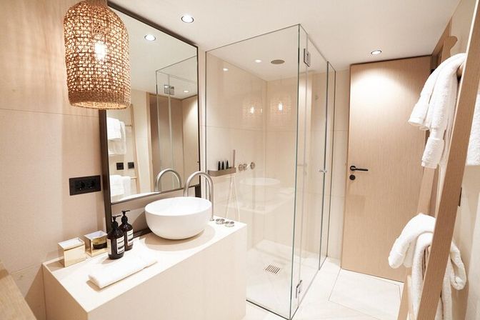 Bright spa-style modern bathroom with glass shower enclosure, white vessel sink on a minimal beige vanity, woven pendant light and neatly folded towels