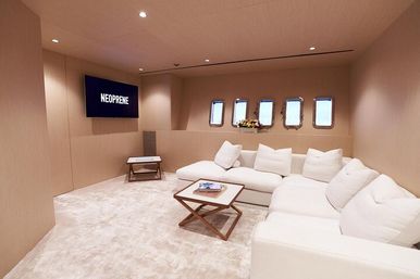 Cozy luxury yacht lounge with cream walls, plush white sectional sofa, wooden coffee tables, a row of porthole windows overlooking the sea, and a wall-mounted TV displaying NEOPRENE.