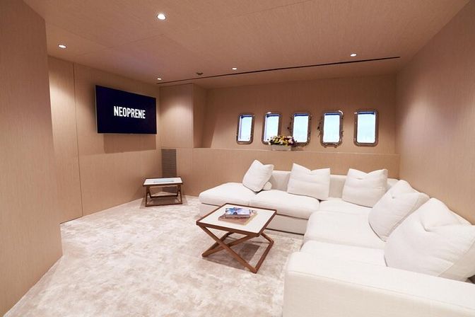 Cozy luxury yacht lounge with cream walls, plush white sectional sofa, wooden coffee tables, a row of porthole windows overlooking the sea, and a wall-mounted TV displaying NEOPRENE.