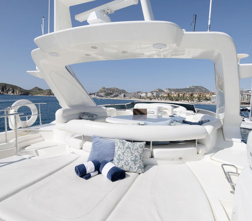 Luxury white yacht deck with plush sun loungers, patterned throw pillows and rolled navy towels, overlooking a calm blue sea and coastal marina under a clear sky.