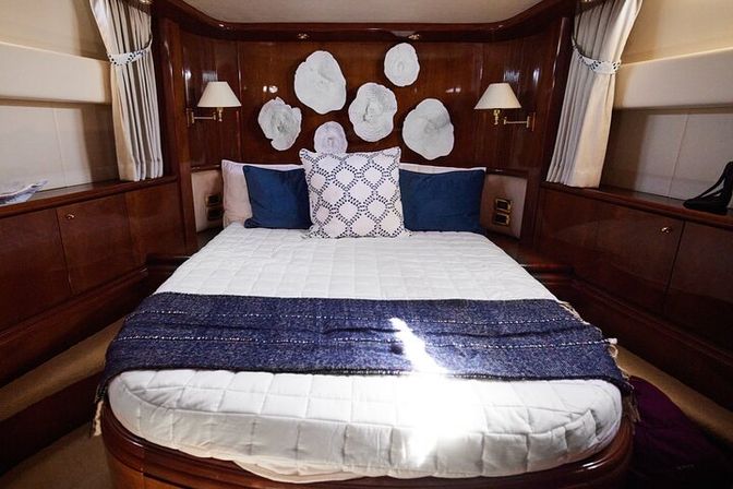 Cozy luxury yacht stateroom with queen bed dressed in white quilt and navy throw, polished wood paneling, blue accent pillows, wall-mounted lamps and white coral-inspired wall art