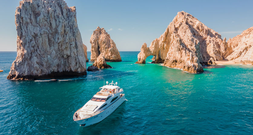 White luxury yacht cruising turquoise waters past dramatic sandstone arches and sea stacks at Los Arcos, Cabo San Lucas on a sunny day