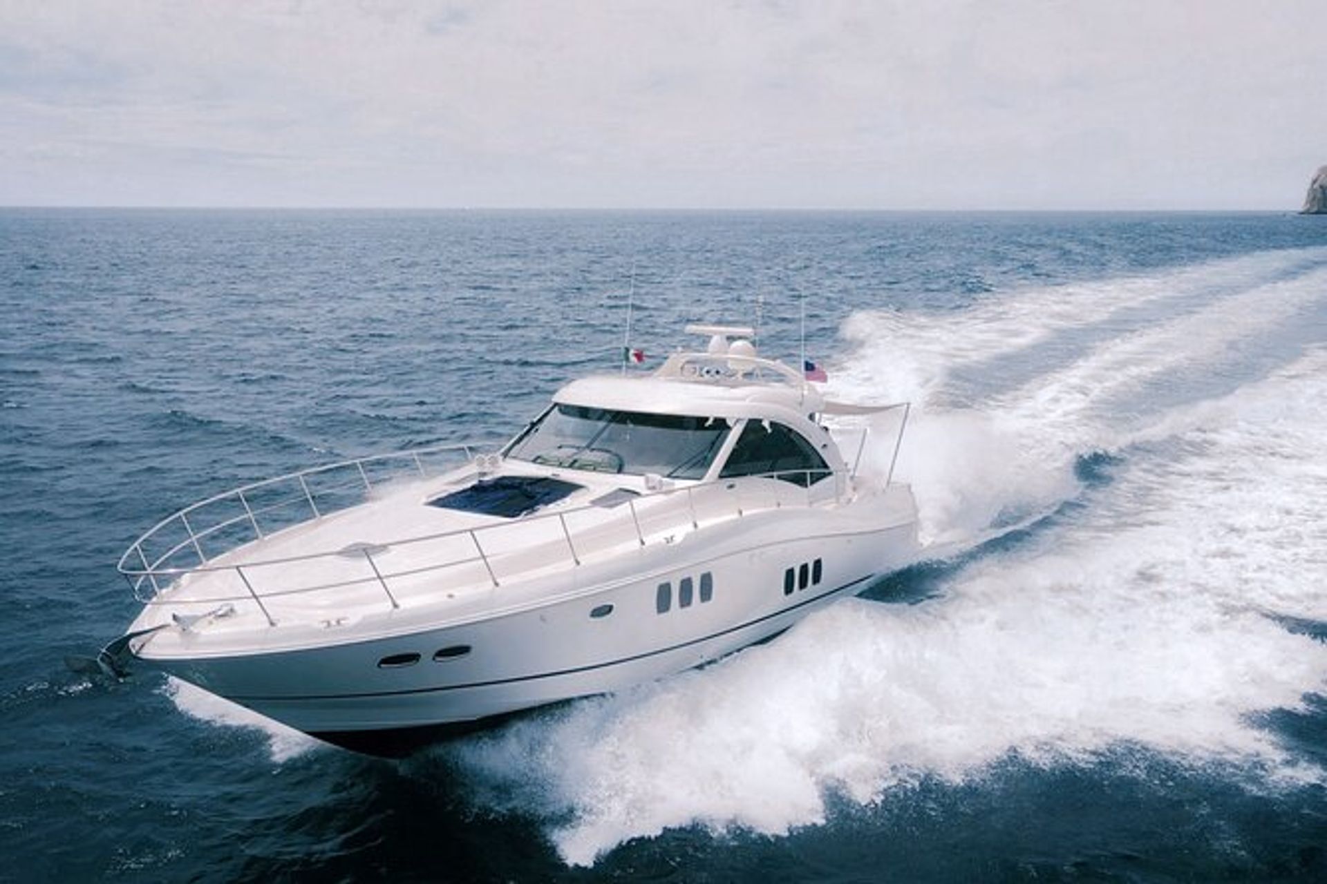 Sleek white luxury motor yacht speeding across deep-blue ocean, cutting a foamy wake under a pale sky with a distant rocky shoreline.