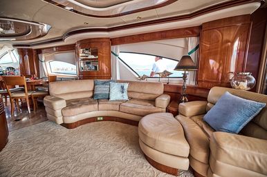 Sunlit luxury yacht salon with curved beige leather sofa and ottoman, polished wood-paneled interior, dining area, decorative pillows and lamp, and large windows framing ocean views.