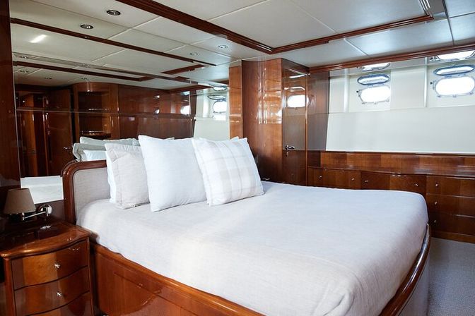 Wood‑paneled luxury yacht stateroom with queen bed in white linens, decorative pillows, glossy built‑in cabinetry and round porthole windows