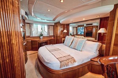 Luxury yacht master stateroom interior with curved wood-paneled walls, plush bed dressed in white linens and decorative pillows, built-in desk, porthole windows, mirrors and warm recessed lighting.