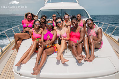 Smiling group of women in vibrant pink swimwear and sunglasses lounging on the bow of a white yacht under sunny blue skies on the open ocean