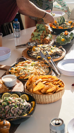 Sunlit buffet spread of tortilla chips, guacamole, nachos, sizzling fajitas and stuffed rolls, with a hand reaching to serve.