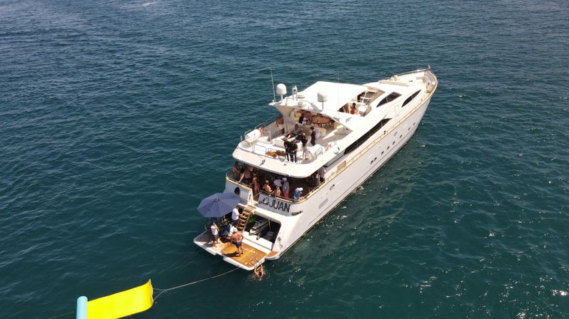 White luxury yacht in deep blue ocean on a sunny day, a group relaxing on multiple decks with an umbrella on the swim platform and a bright yellow inflatable water slide tethered to the stern.