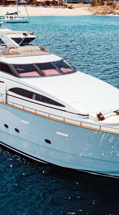 Sleek white luxury yacht cruising clear blue water near a sandy beach with other boats in the background.