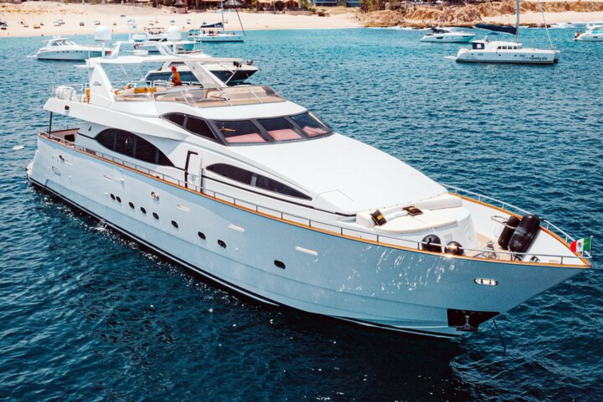 Sleek white luxury yacht cruising clear blue water near a sandy beach with other boats in the background.