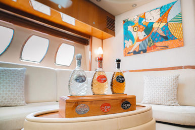 Luxury yacht interior lounge with three decorative liquor bottles on a wooden box atop a round ottoman, cream leather seating, porthole windows and vibrant abstract wall art.
