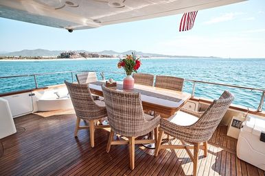 Luxury yacht teak deck with wicker dining table and chairs, pink vase of flowers, turquoise ocean and distant coastal hills under a sunny sky.