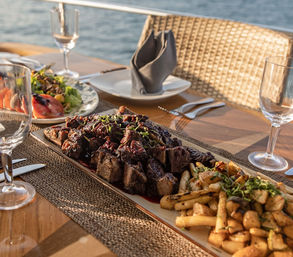 Waterfront outdoor dining: mouthwatering grilled steak bites on a long platter with roasted potatoes, mixed salad, wine glasses and a wicker chair at sunset.