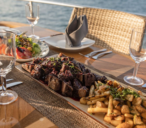 Waterfront outdoor dining: mouthwatering grilled steak bites on a long platter with roasted potatoes, mixed salad, wine glasses and a wicker chair at sunset.