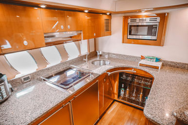 Luxury yacht galley kitchen with glossy wood cabinetry, granite countertops, electric cooktop, microwave, round sink, porthole windows and glass-front liquor cabinet