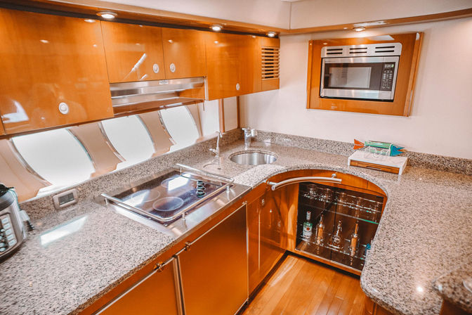 Luxury yacht galley kitchen with glossy wood cabinetry, granite countertops, electric cooktop, microwave, round sink, porthole windows and glass-front liquor cabinet