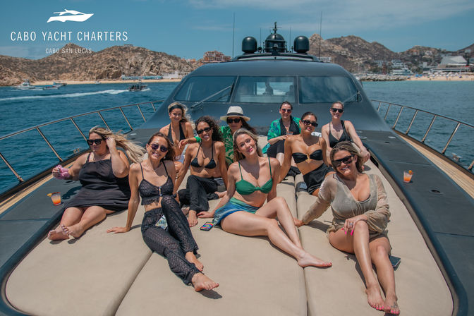 Group of women sunbathing and smiling on the foredeck of a luxury yacht off Cabo San Lucas with rocky coastline and blue ocean