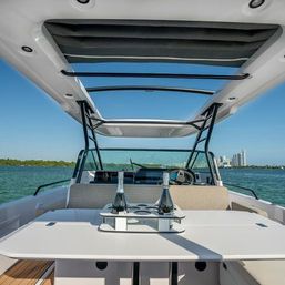 Luxury motorboat cockpit with open hardtop and sunshade, table holding two champagne bottles, turquoise coastal water and distant city skyline under a clear blue sky