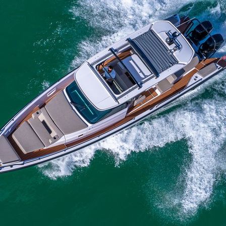 Aerial shot of a sleek luxury speedboat with teak accents and twin outboard engines cutting through emerald-green water, leaving a foamy white wake as a person steers under a shaded hardtop.