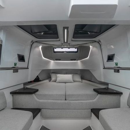Sleek modern yacht cabin with a double forward berth, grey cushions and pillows, overhead skylights, LED accent lighting and built-in side tables and seating — cozy minimalist marine interior.