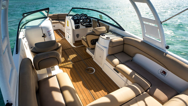 Sunlit luxury boat interior with teak-style deck, tan cushioned wraparound seating, swivel captain’s chair and helm console, open windshield overlooking clear turquoise water.