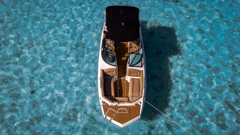Aerial view of a white motorboat with tan seating and teak deck anchored in crystal-clear turquoise water, sunlit tropical scene