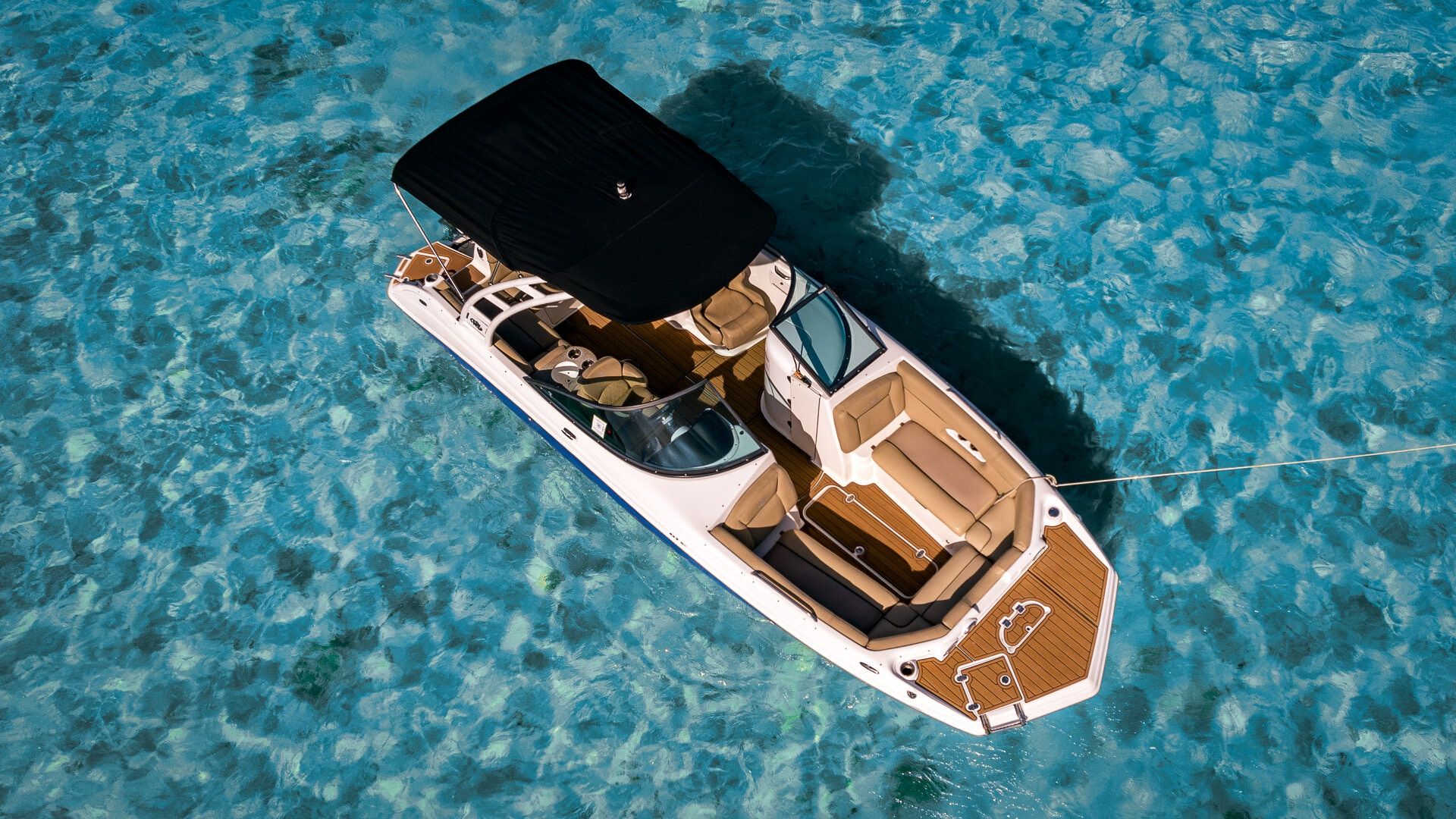 Aerial shot of a white motorboat with tan cushions and black canopy moored by a rope over crystal-clear turquoise tropical water, sunlight revealing the sandy seabed.