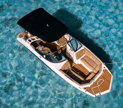 Aerial shot of a white motorboat with tan cushions and black canopy moored by a rope over crystal-clear turquoise tropical water, sunlight revealing the sandy seabed.
