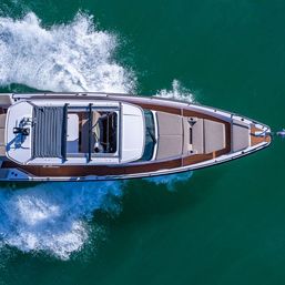 Aerial drone view of a sleek luxury motorboat cruising through turquoise water, leaving a foamy white wake alongside its teak deck and sunbed area.