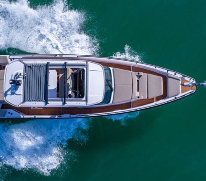 Aerial drone view of a sleek luxury motorboat cruising through turquoise water, leaving a foamy white wake alongside its teak deck and sunbed area.