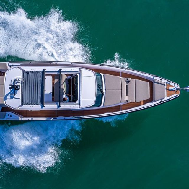 Aerial drone view of a sleek luxury motorboat cruising through turquoise water, leaving a foamy white wake alongside its teak deck and sunbed area.