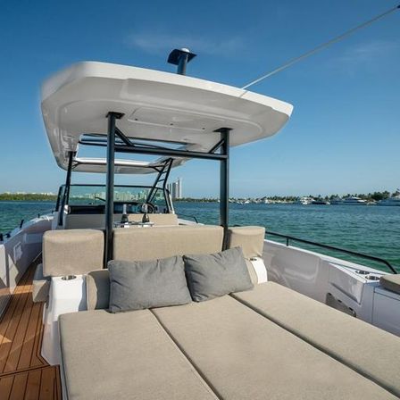 Luxury motorboat sunbed with beige cushions and gray pillows under a T-top canopy, overlooking a turquoise bay and distant marina under a clear blue sky