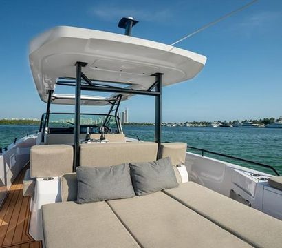 Luxury motorboat sunbed with beige cushions and gray pillows under a T-top canopy, overlooking a turquoise bay and distant marina under a clear blue sky